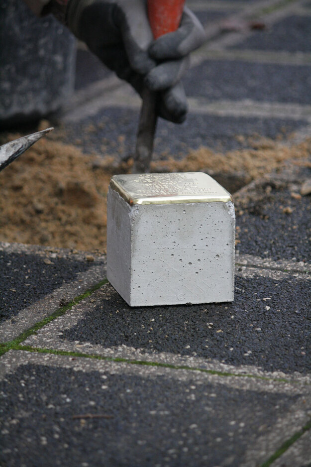 Detailaufnahme von einem Stolperstein kurz vor der Verlegung, er liegt auf dem Boden, im Hintergrund wird die Lücke in den Pflastersteinen vorbereitet