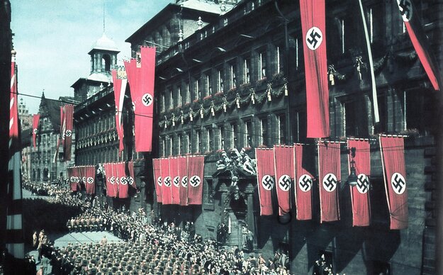 Kolorierte Fotografie vom Nürnberger Rathaus, an dessen Fassade rote Hakenkreuzflaggen wehen, in der Straße vor dem Gebäude marschieren Nationalsozialisten