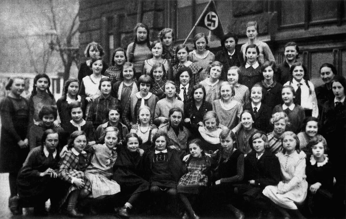 Klassenfoto der Schülerinnen der ehemaligen Städtischen Höheren Mädchenschule in Nürnberg, April 1933. Sammlung Frank Harris, New York. Schwarz-weiß Fotografie auf der eine Gruppe Schülerinnen abgebildet sind. Im Hintergrund ist ein Gebäude.